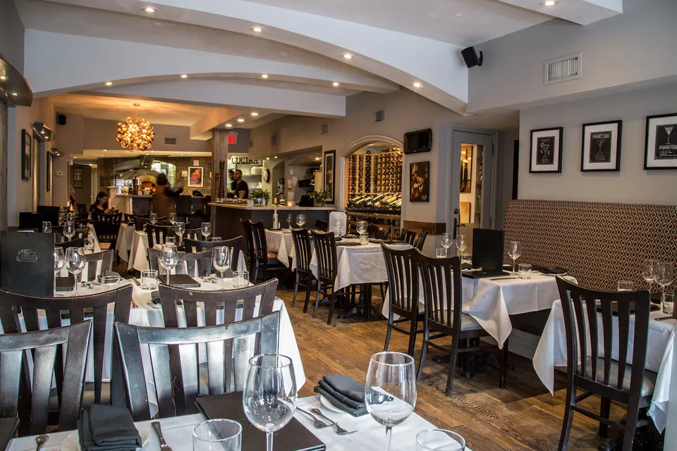 An elegant dining room at Trio Ristorante Pizzeria featuring dark wood chairs, white tablecloths, set wine glasses, and arched ceiling beams leading to a bar area.