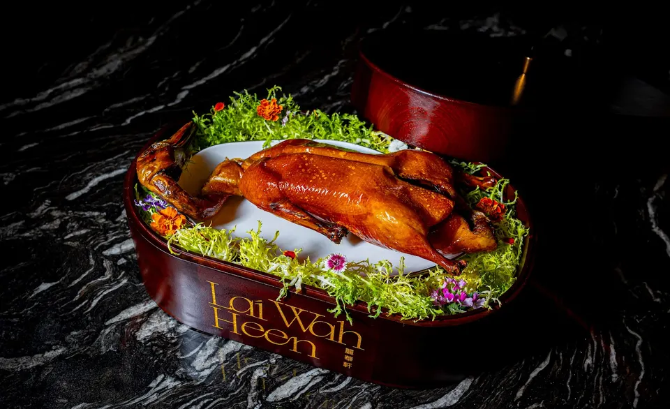 A whole roasted Peking duck with glossy, golden-brown skin, served on a white platter lined with fresh greens and edible flowers inside a dark wooden box labeled Lai Wah Heen.