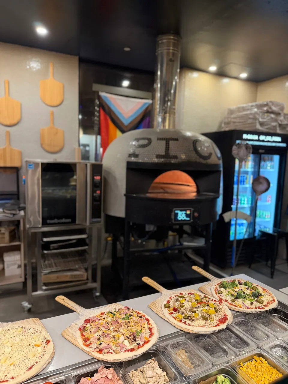 Four uncooked pizzas on wooden peels sit on a prep counter in front of a large, dome-shaped stone pizza oven with a digital display.