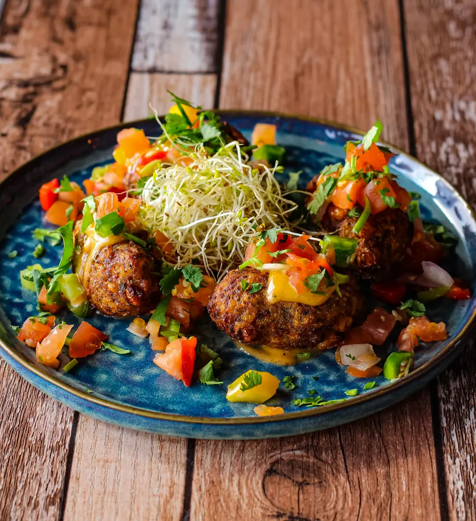 Four golden-brown fritters arranged on a blue ceramic plate, topped with melted cheese, fresh tomato salsa, chopped cilantro, and a mound of alfalfa sprouts in the center.