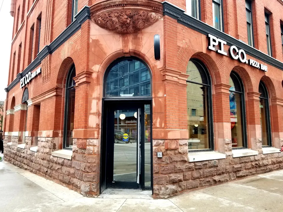 Exterior of Pi Co. Pizza Bar located in a historic red brick building with arched windows and stone foundation on a city street corner.