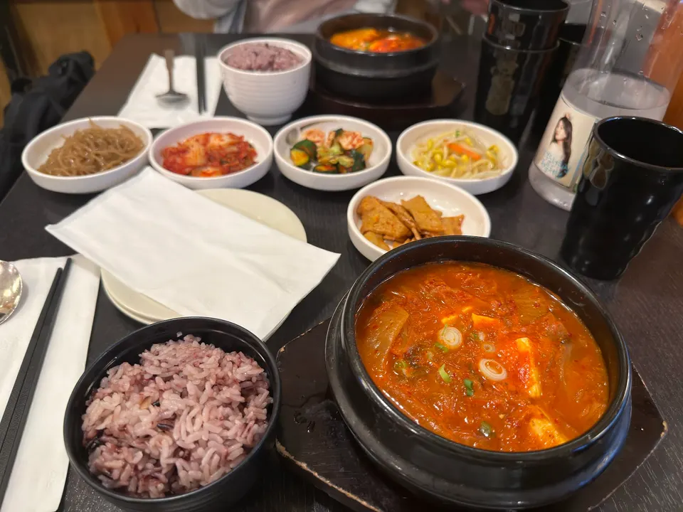 A table set with Korean dishes including two bowls of purple rice, a large stone pot of bubbling red kimchi stew, and several small side dishes like kimchi, bean sprouts, and fish cakes.