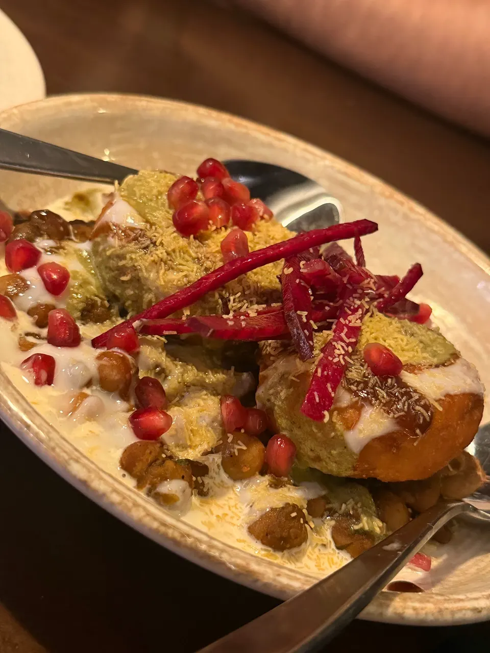 A close-up shot of a plate of Dahi Bhalla chaat, topped with yogurt, green chutney, sweet tamarind chutney, sev, pomegranate seeds, and julienned beetroot, served with spoons.