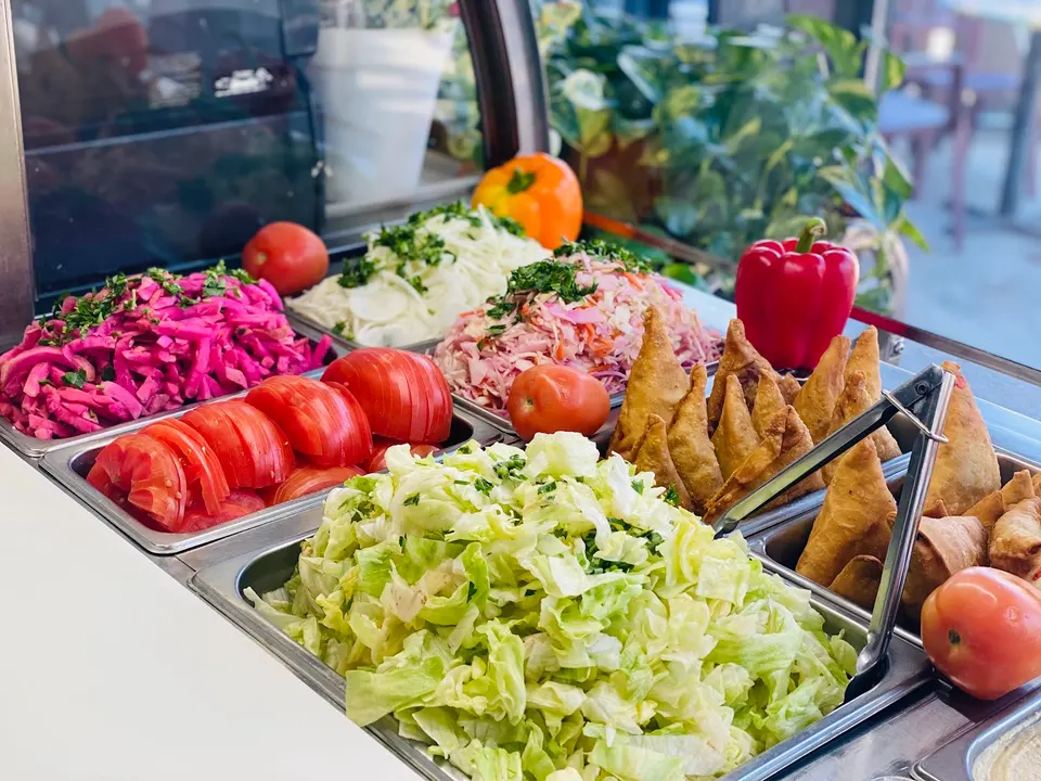 A metal food display counter filled with fresh salad ingredients including shredded lettuce, sliced tomatoes, pickled red cabbage, onions, and golden-brown samosas.