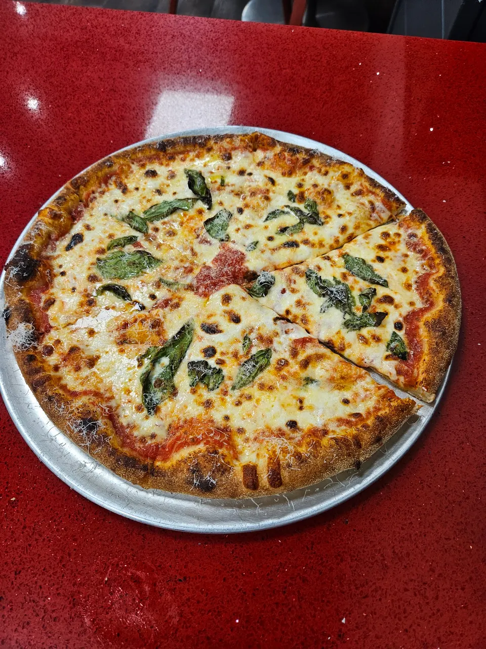 A freshly baked Margherita pizza with melted mozzarella cheese, tomato sauce, and fresh basil leaves, served on a metal tray on a red countertop.