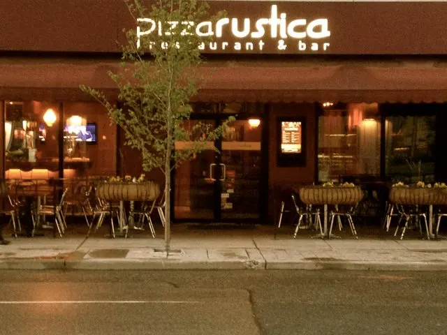 The exterior storefront of Pizza Rustica restaurant and bar at night, featuring outdoor patio seating with tables and chairs, a small tree, and a glowing sign above the entrance.