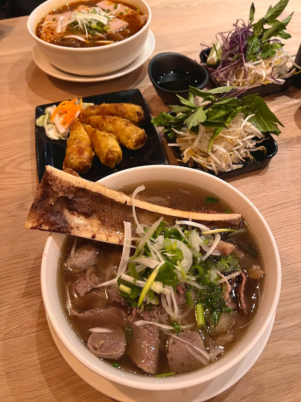 A table spread at a Vietnamese restaurant featuring a large bowl of beef pho with a roasted bone marrow bone, a bowl of spicy noodle soup, fried spring rolls, and fresh herb garnishes.
