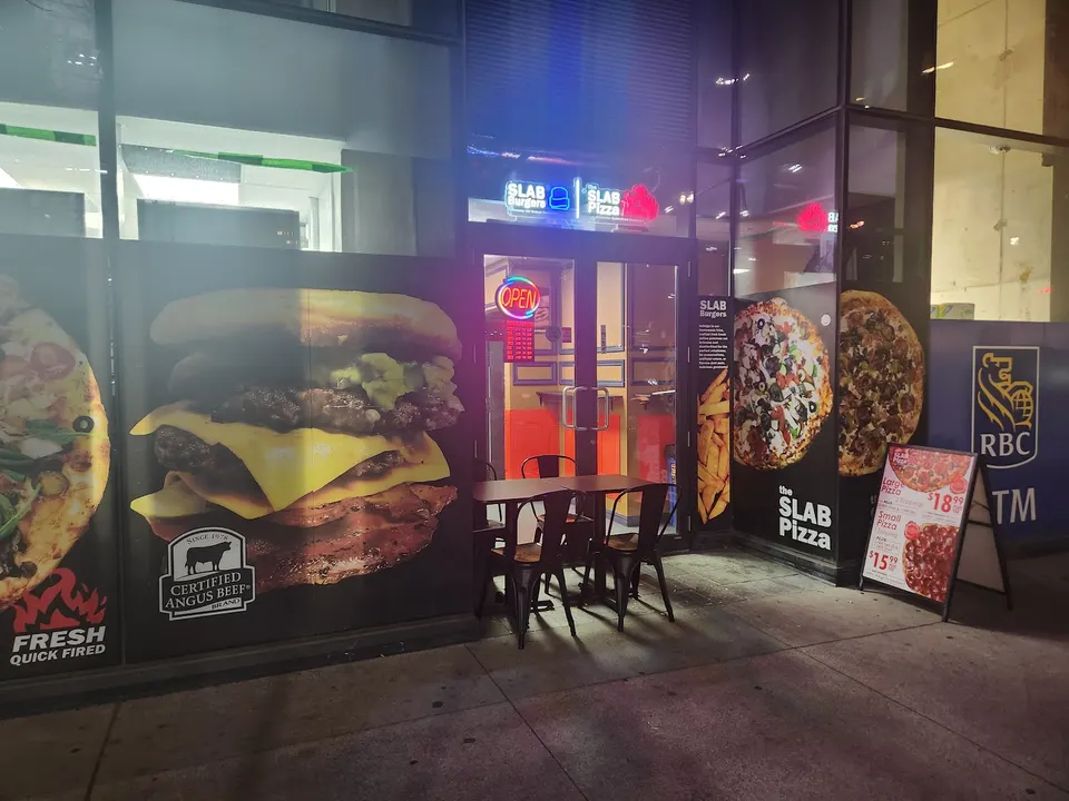 Exterior storefront of The Slab Pizza at night, featuring large window graphics of a double cheeseburger and pizzas, with outdoor seating and an open sign glowing in the doorway.