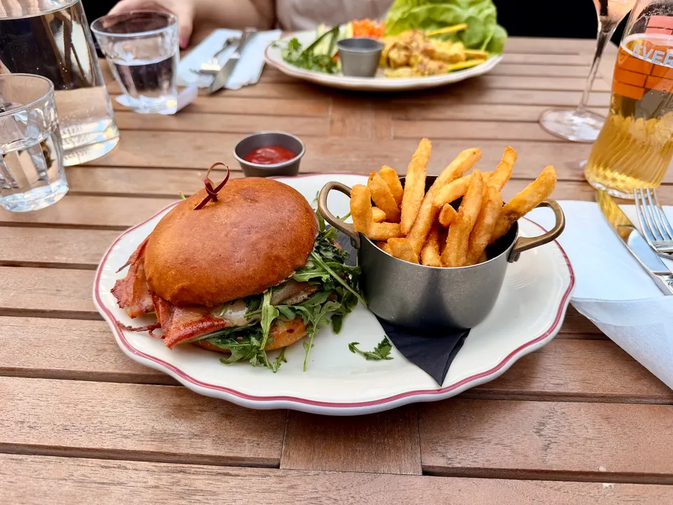 A gourmet burger with bacon and arugula on a brioche bun, served on a white plate with a side of golden french fries in a small metal cup, on an outdoor wooden table.
