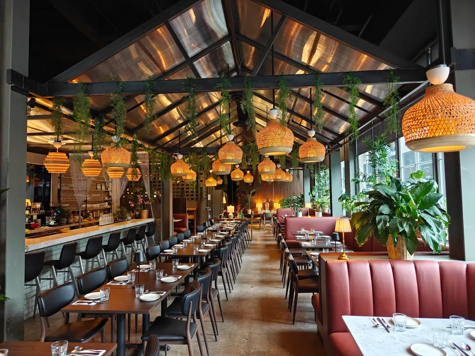 A spacious restaurant interior with long wooden tables, red leather booths, bar seating, hanging woven pendant lights, and lush green plants under a glass greenhouse-style roof.
