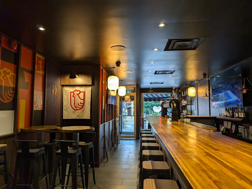 Interior of Tebasaki Wing House featuring a long wooden bar counter with stools, high-top tables with metal chairs, Japanese-style lanterns, and a rooster logo banner on the wall.