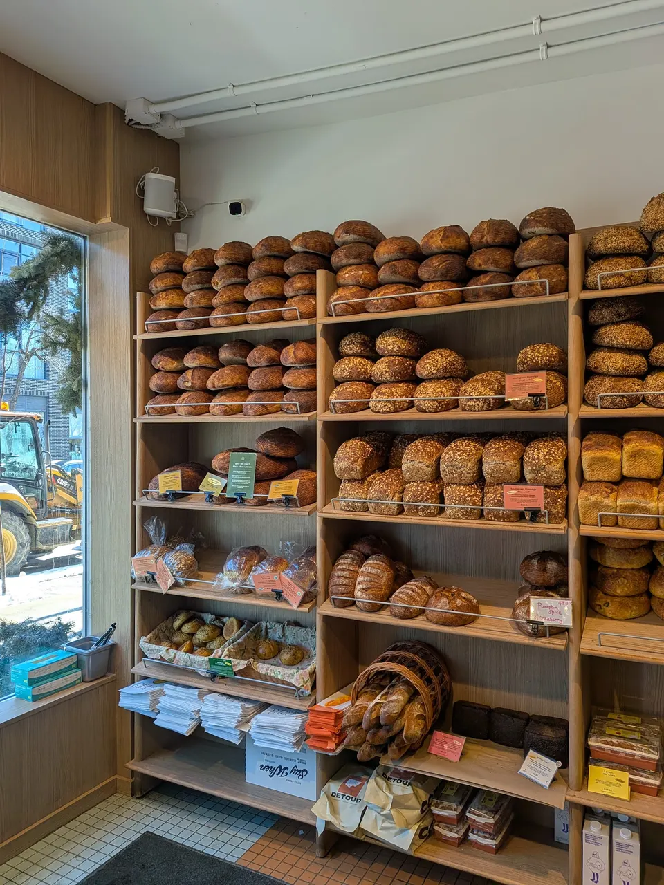 Wooden shelves filled with various freshly baked artisanal bread loaves, including sourdough, seeded, and baguettes, inside Dear Grain bakery.
