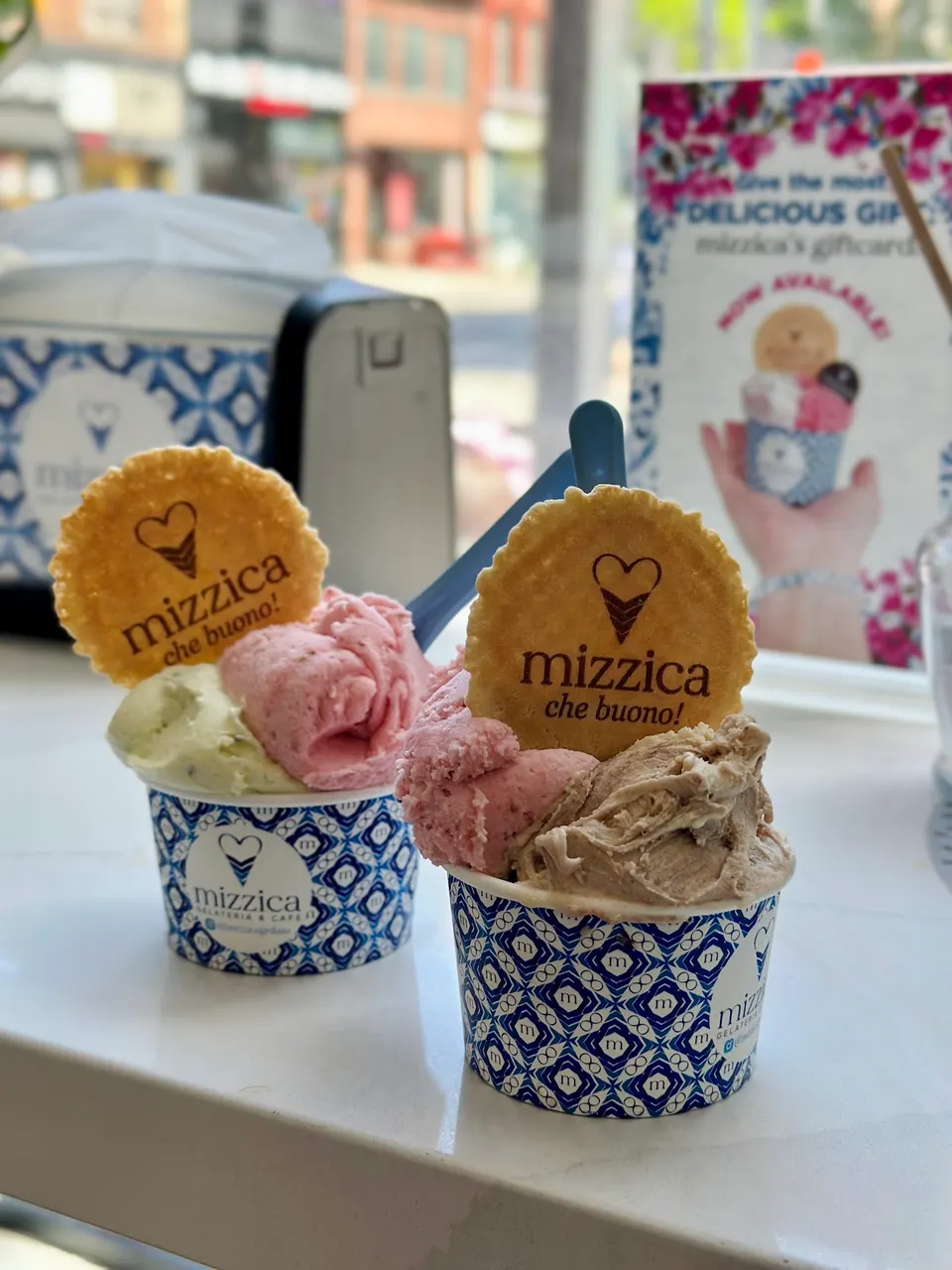 Two cups of artisanal gelato on a white counter at Mizzica Gelateria & Cafe. One cup has pistachio and strawberry gelato, the other has strawberry and hazelnut gelato, both topped with a round waffle cookie featuring the shop's logo.