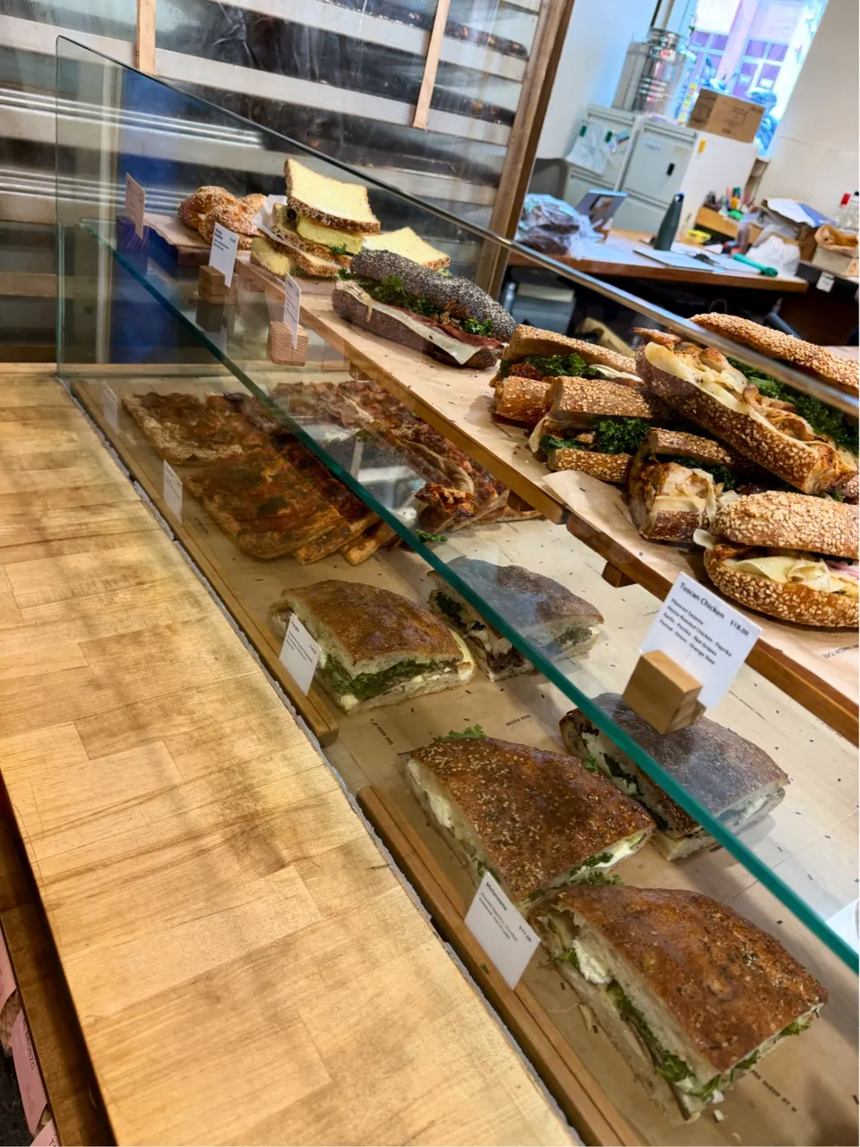 A glass display case at Forno Cultura filled with various gourmet sandwiches, including focaccia sandwiches, seeded baguettes, and sliced paninis on wooden shelves.