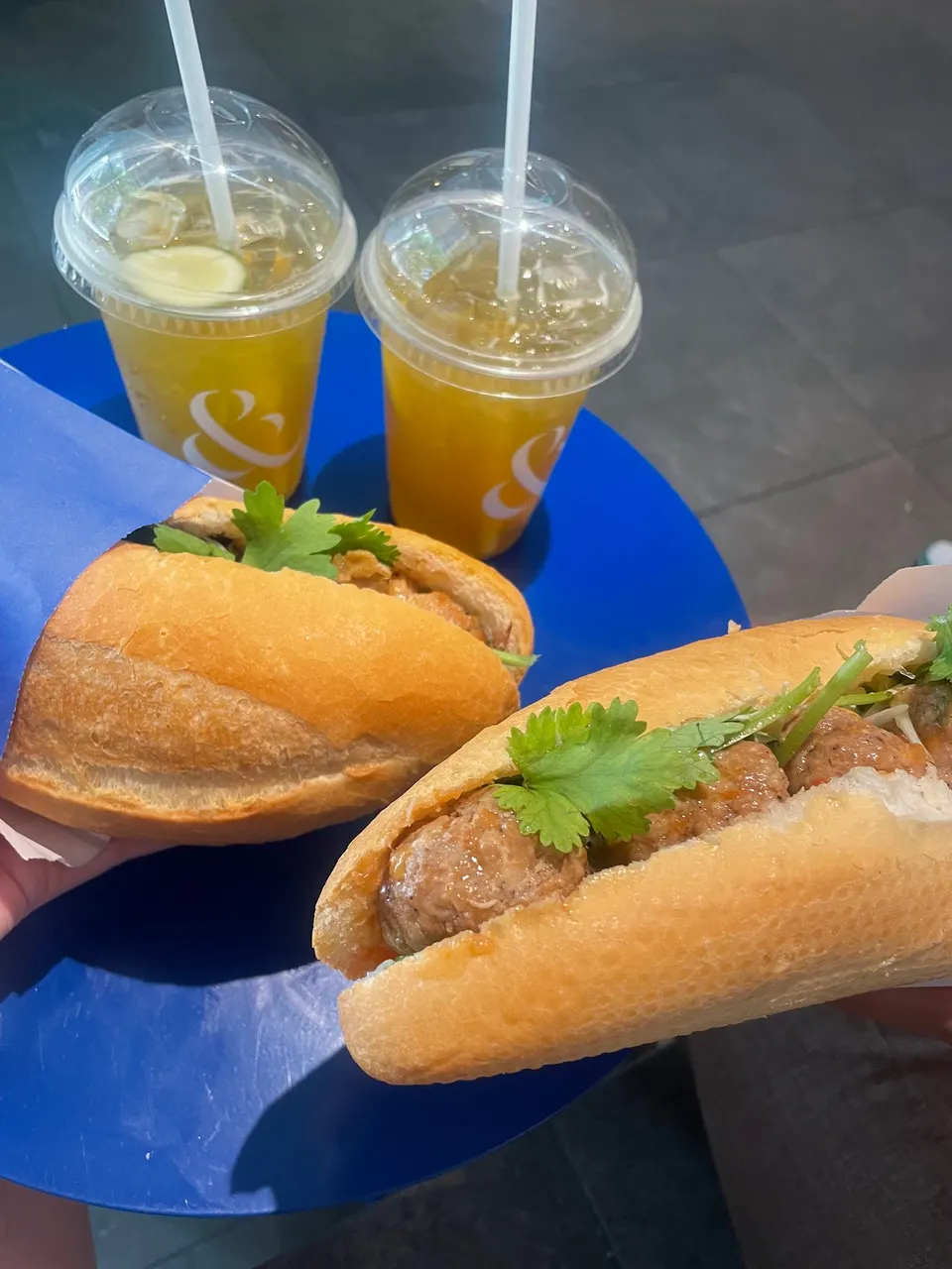 Two Vietnamese banh mi sandwiches filled with meatballs and fresh cilantro, held in hand over a blue table next to two iced tea drinks in plastic cups.