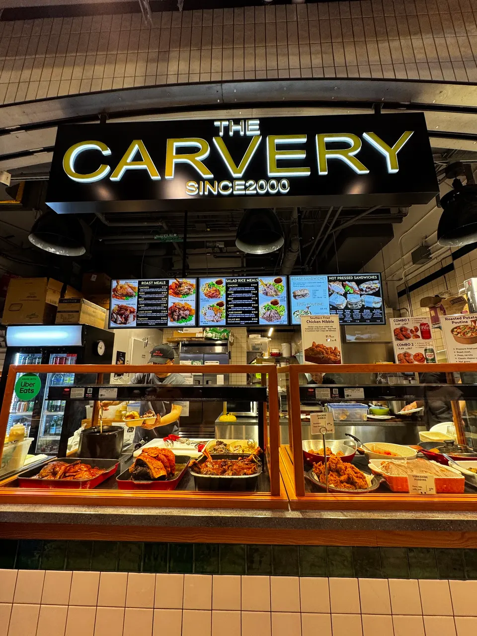 The storefront of The Carvery at The Well food court, featuring a glowing yellow sign, overhead menus, and a glass display case filled with roasted meats and hot side dishes.