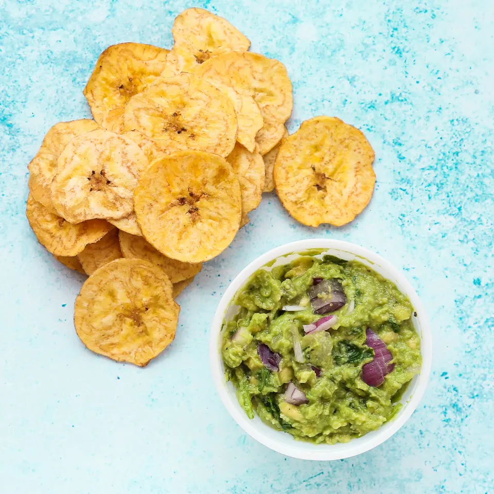 A top-down view of crispy, golden plantain chips piled next to a small white bowl filled with fresh guacamole containing visible red onion chunks, set against a textured light blue background.