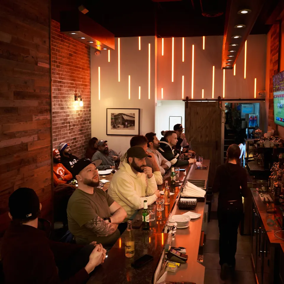 Patrons sitting along a long bar counter watching a screen in a dimly lit bar with brick walls and vertical light strips on the back wall.