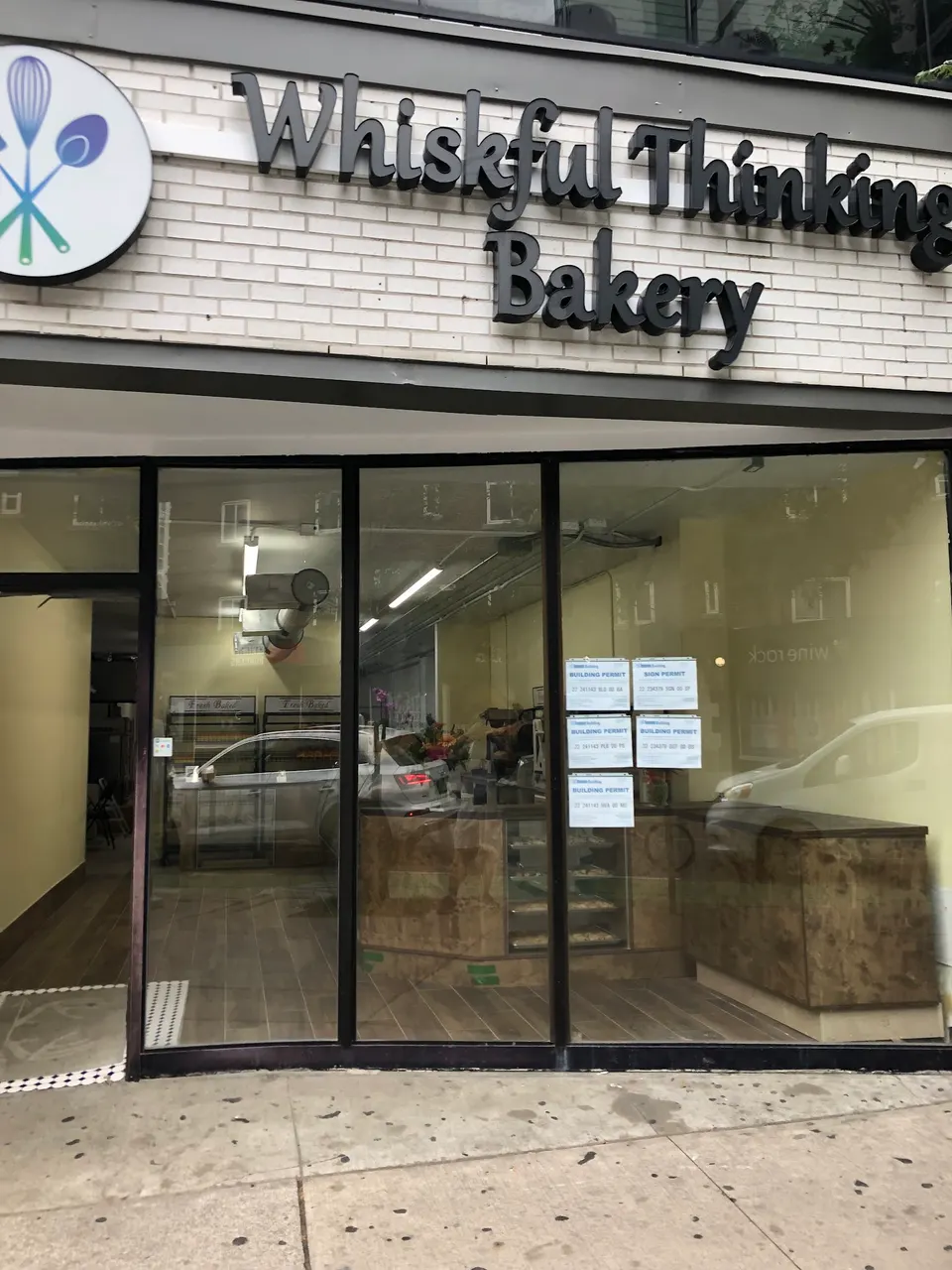 The storefront of Whiskful Thinking Bakery, featuring a white brick facade, a black sign with the bakery's name, and large glass windows showing a counter inside.