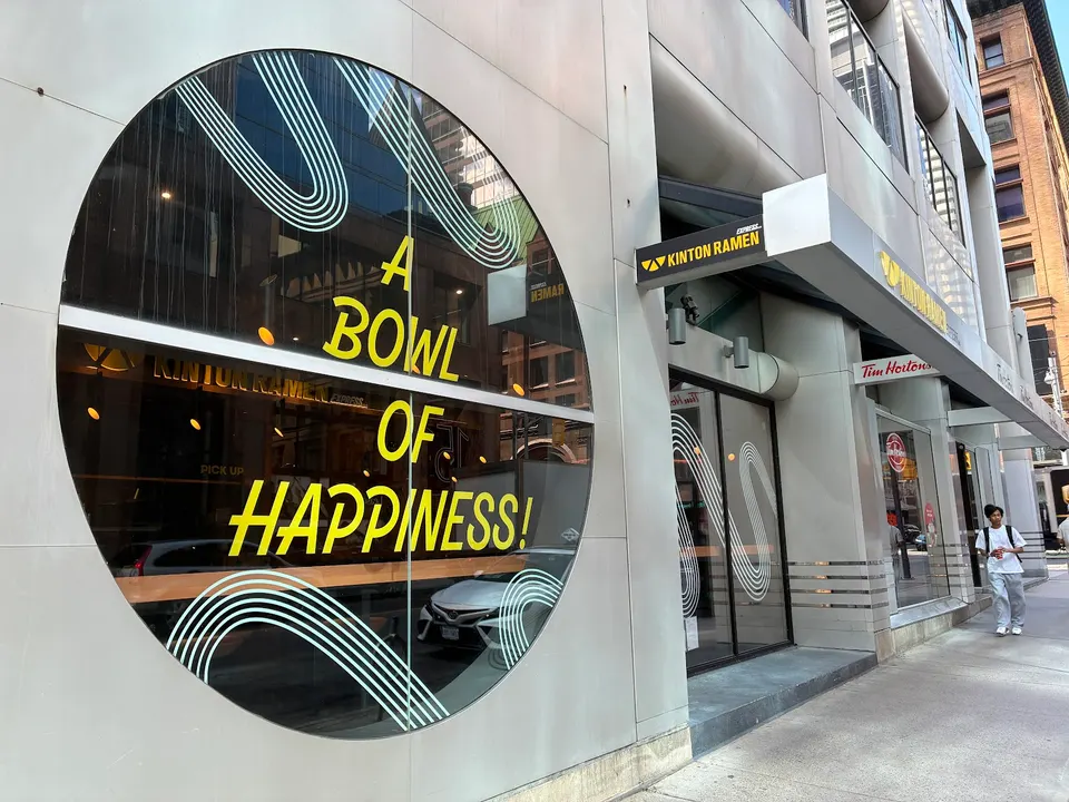 The storefront of Kinton Ramen on Yonge and Richmond, featuring a large circular window with the yellow text 'A BOWL OF HAPPINESS!' and the restaurant sign above the entrance.