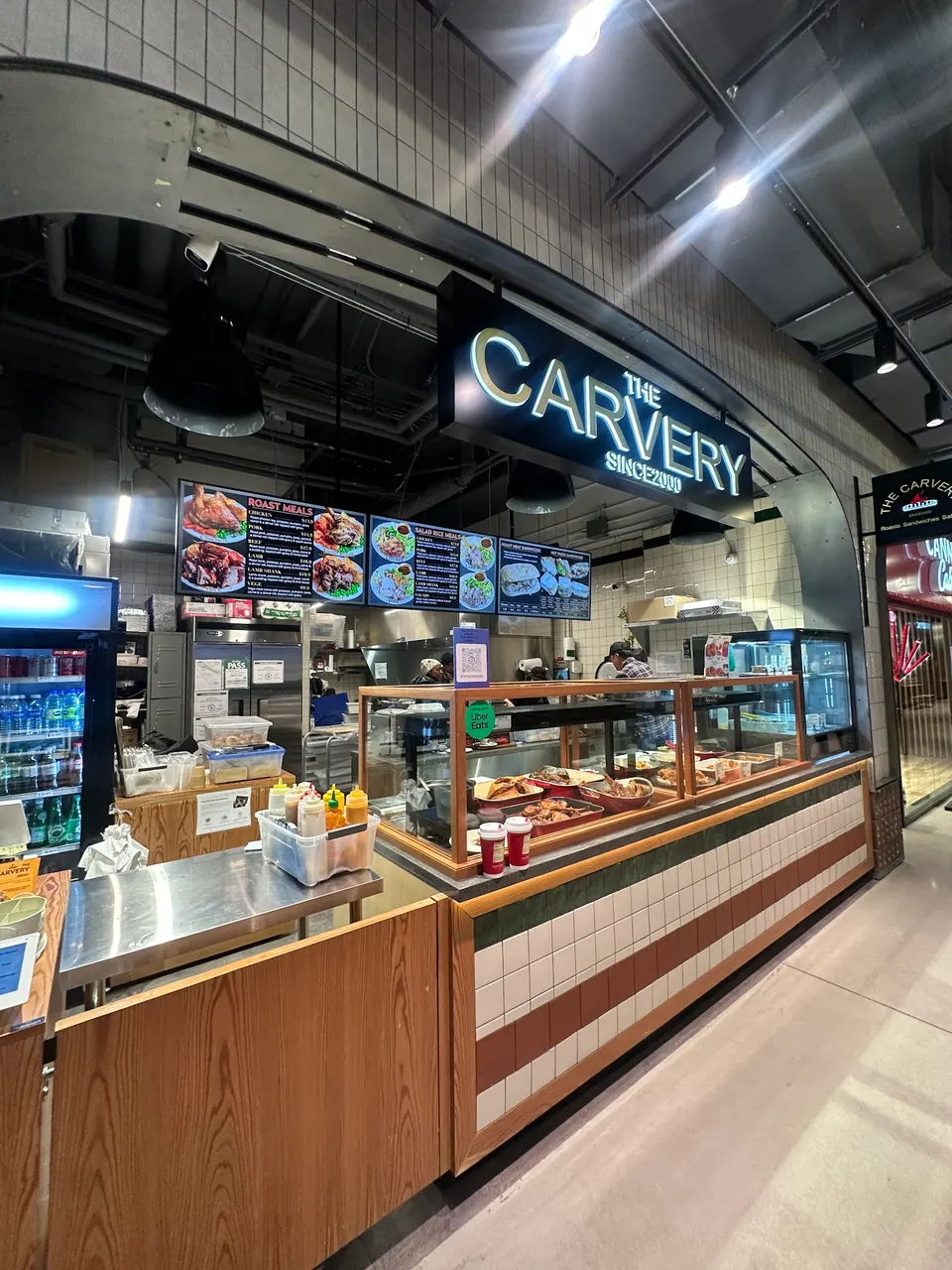 A modern food court stall named The Carvery, featuring a glass display case with roasted meats, a menu board overhead, and a clean wooden counter.