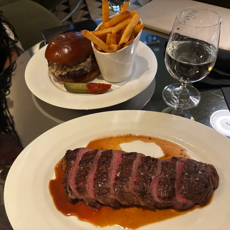 A sliced medium-rare steak with jus on a white plate in the foreground, with a burger, french fries, and a glass of water on a reflective table in the background.