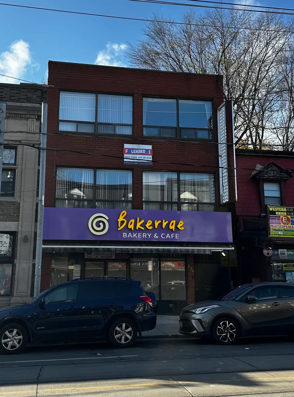 Exterior view of Bakerrae Bakery & Cafe, featuring a purple storefront sign with a yellow swirl logo and text, set on a brick building with large windows and cars parked in front.