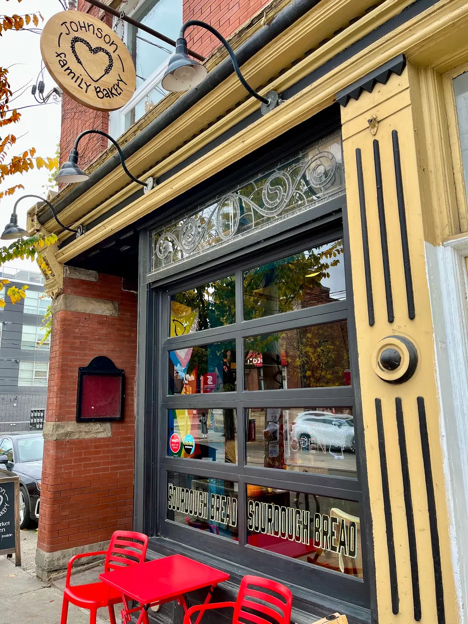 Exterior of Johnson Family Bakery featuring a wooden circular sign, a large glass garage-style window with 'SOURDOUGH BREAD' lettering, and a small red outdoor table with two red chairs.
