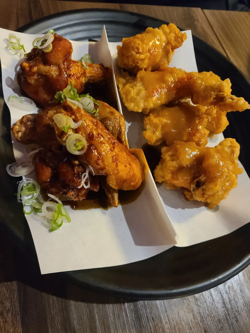 A black plate containing two varieties of fried chicken wings served on white paper liners. One side features wings coated in a dark, glossy sauce topped with sliced green onions, while the other side has golden-brown wings drizzled with a creamy yellow sauce.