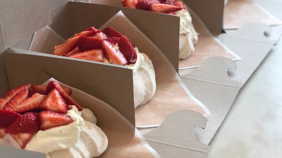 Three individual strawberry pavlova desserts, each with a meringue base, whipped cream, and fresh sliced strawberries, arranged in individual cardboard takeout boxes.