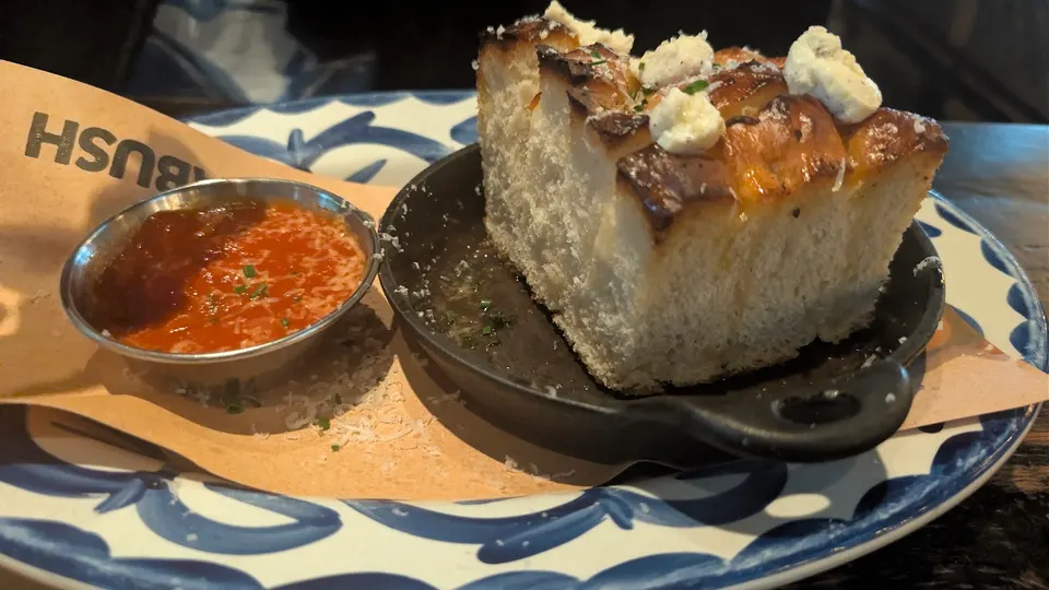 A thick slice of toasted focaccia bread topped with whipped cheese, served in a small cast-iron skillet next to a small metal cup of marinara sauce on a blue and white patterned plate.