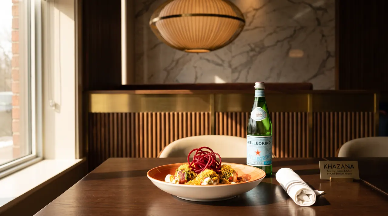 A beautifully plated Indian street food dish topped with bright red spiral garnishes, served alongside a bottle of S.Pellegrino sparkling water on a wooden table at Khazana.