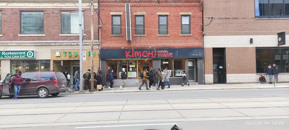 Street view of storefronts including Tsujiri and Kimchi Korea House, with pedestrians walking on the sidewalk and a maroon minivan parked on the street.