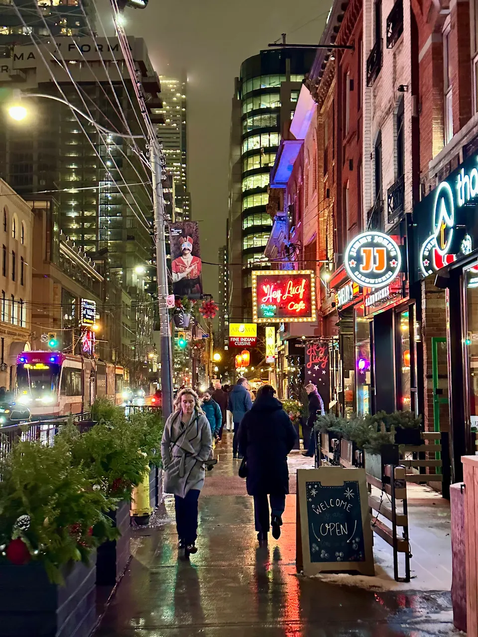 A bustling city sidewalk at night with pedestrians walking past illuminated storefronts, including the glowing red neon sign for Hey Lucy Cafe.