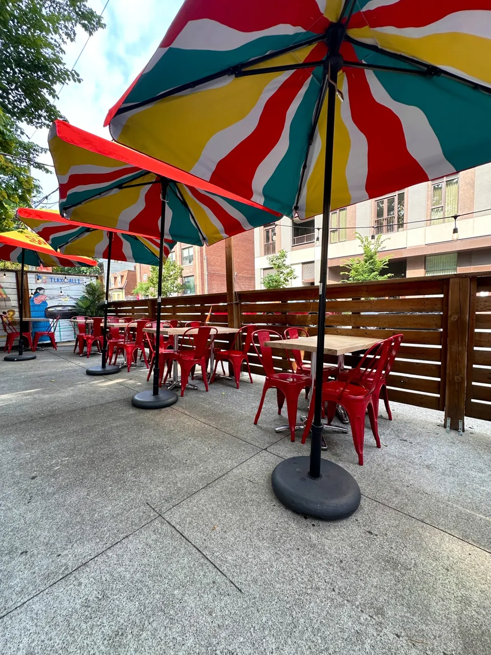 An outdoor patio dining area with concrete flooring, wooden fencing, and several small tables paired with bright red metal chairs. Large, colorful striped umbrellas in red, yellow, teal, and white shade each table.