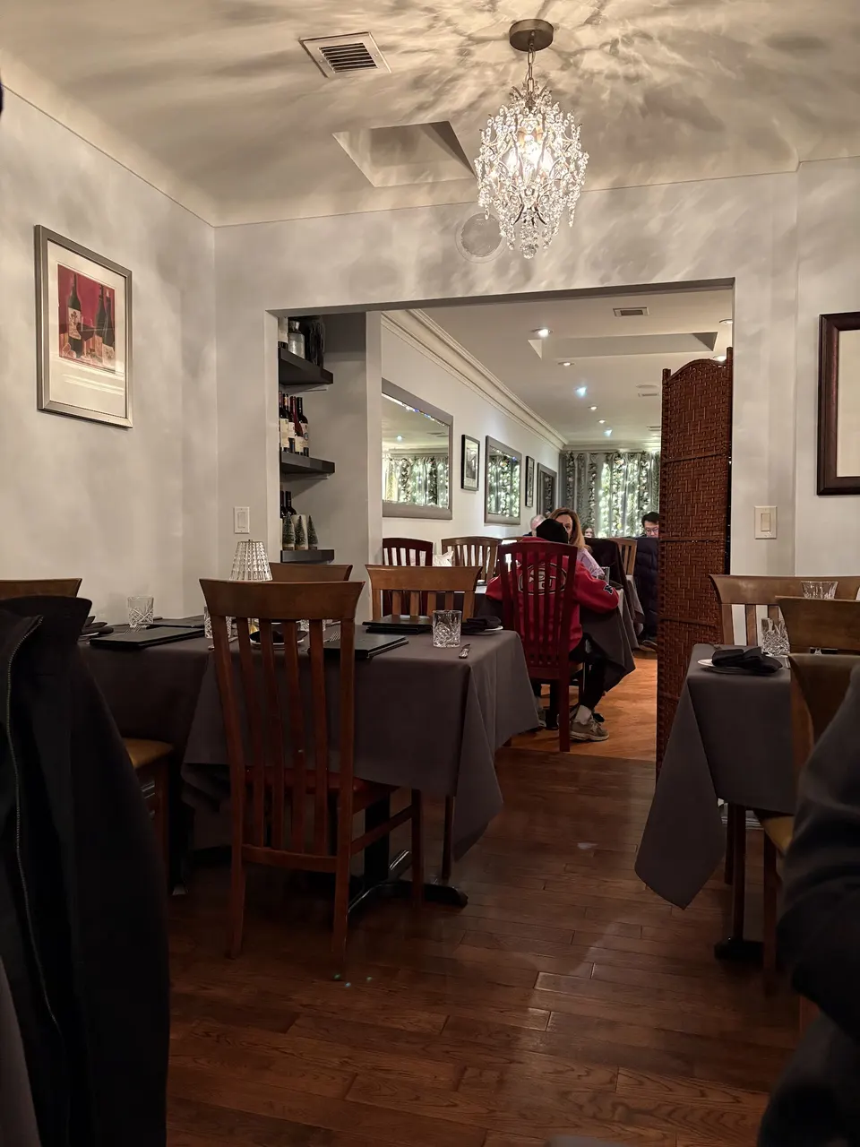 An elegant dining room at Trio Ristorante Pizzeria featuring dark wood chairs, tables set with grey tablecloths, and a crystal chandelier hanging from the ceiling.