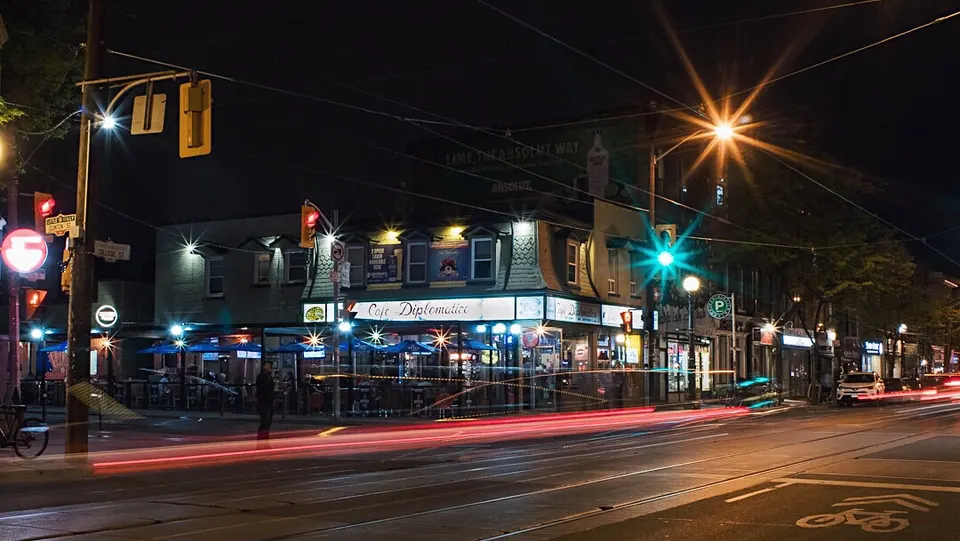 A long exposure night shot of Cafe Diplomatico Restaurant & Pizzeria at a street corner, featuring bright streetlights, glowing signage, and red light trails from passing traffic.