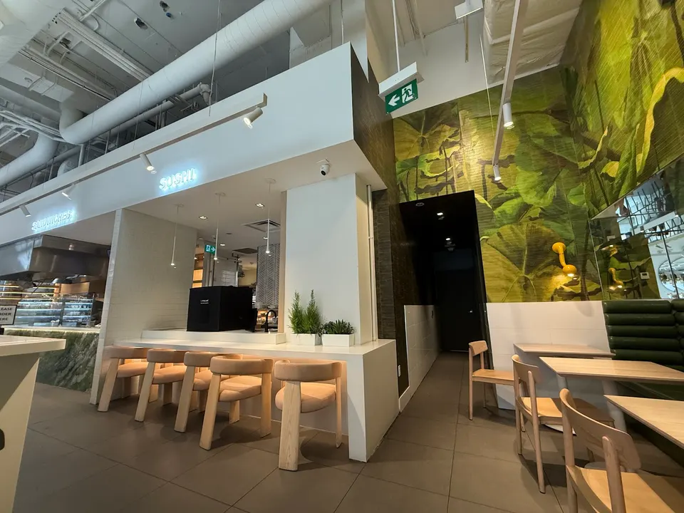 Modern interior of Green Box restaurant featuring a sleek white sushi counter with light wood stools, dining tables with matching chairs, and a vibrant green leaf-patterned accent wall.