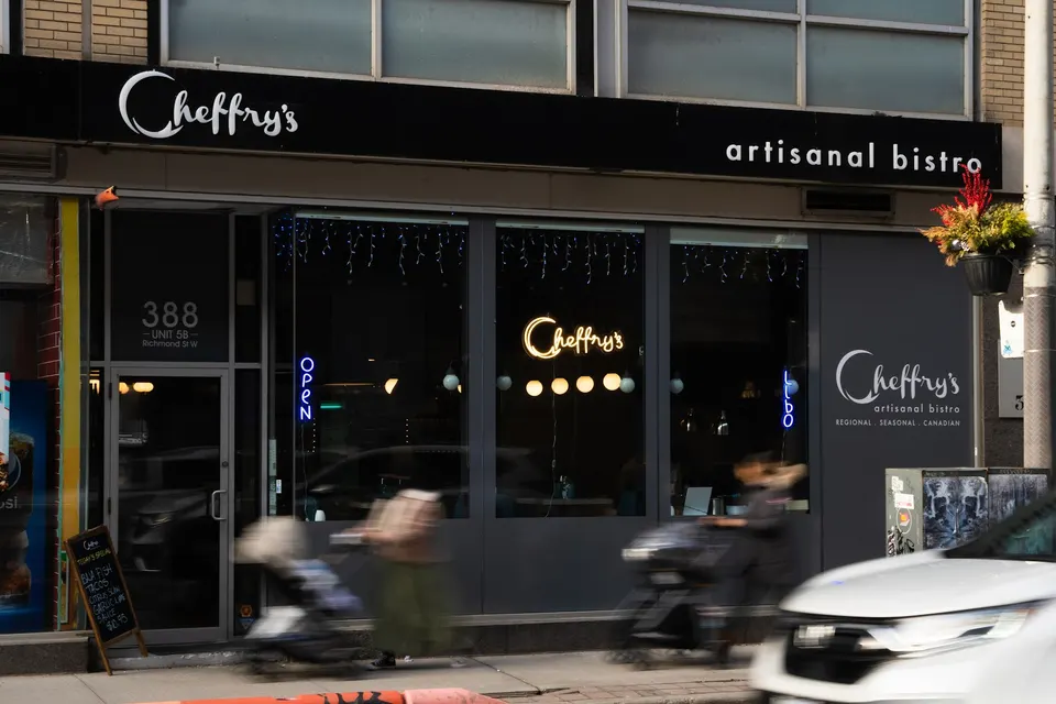 The storefront of Chefry's artisanal bistro at dusk, featuring a dark grey facade, glowing neon signs in the windows, and blurred figures of pedestrians passing by on the sidewalk.