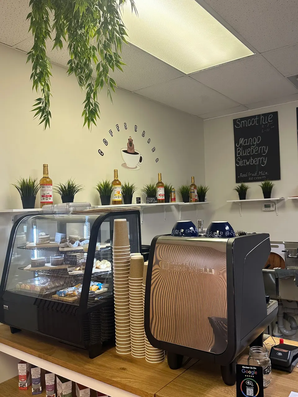A coffee shop counter featuring a glass pastry display case, tall stacks of paper cups, and a professional espresso machine. A menu board on the wall lists smoothie options.