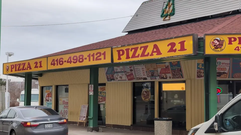 Exterior storefront of Pizza 21 showing its yellow and red sign with phone number 416-498-1121, menu posters on the windows, and a grey sedan parked in front.
