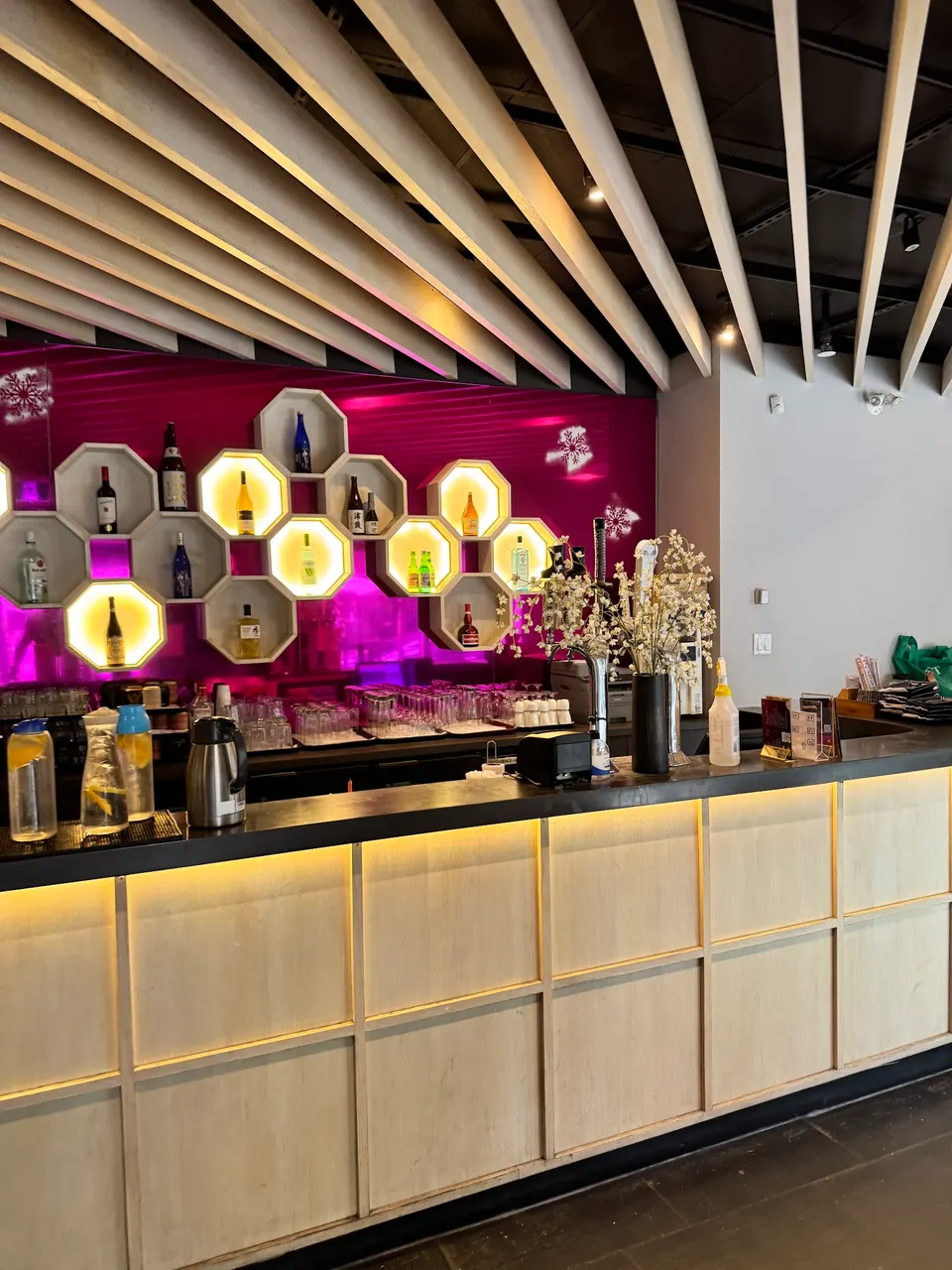 A modern bar counter with light wood paneling and under-counter lighting, featuring a back wall with hexagonal shelves displaying liquor bottles against a vibrant pink background.