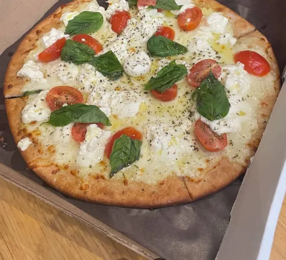 A freshly baked gourmet white pizza topped with melted mozzarella, dollops of fresh burrata cheese, halved cherry tomatoes, and fresh green basil leaves, served in a cardboard delivery box.