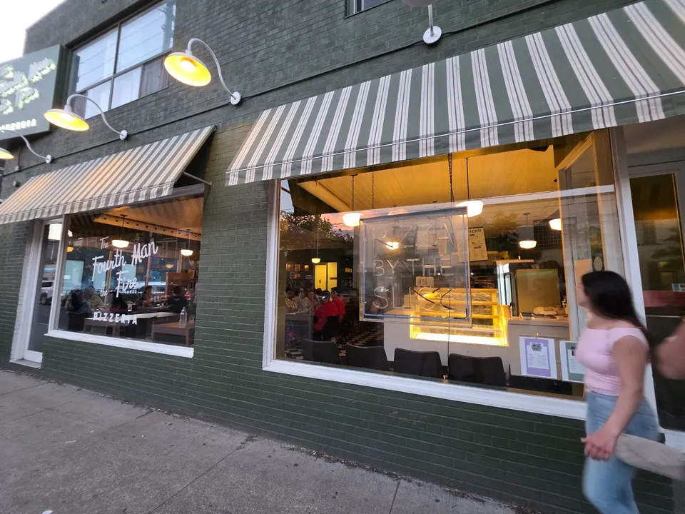 Street view of The Fourth Man in the Fire Pizzeria showing large glass windows, green brick exterior, striped awnings, and warm indoor lighting with a person walking past.