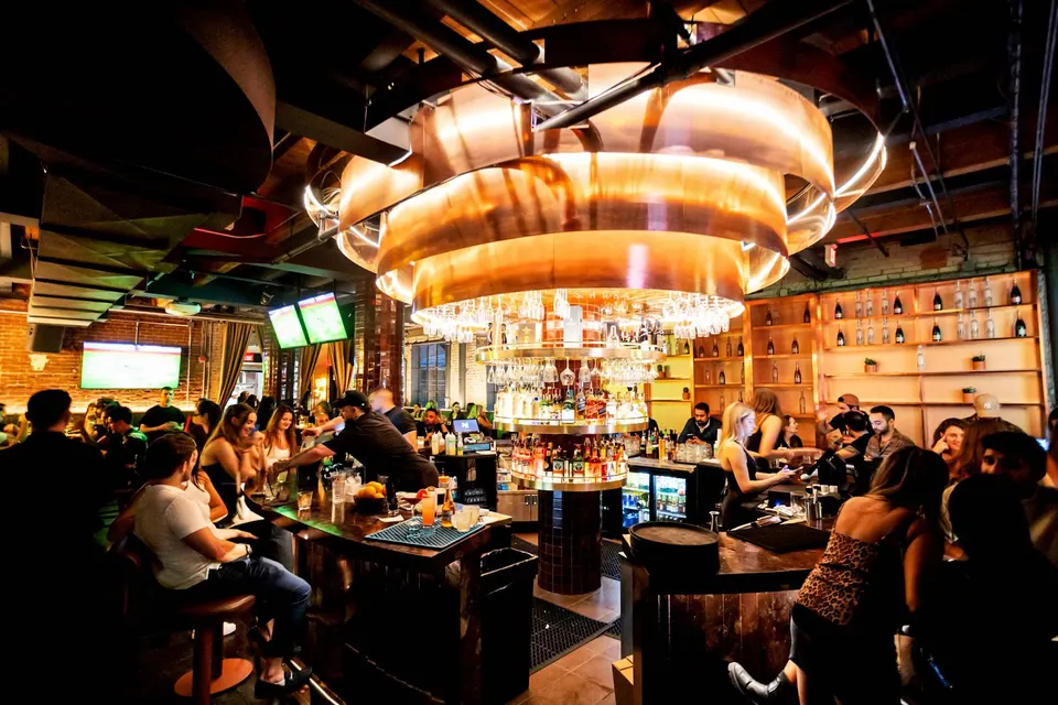 A bustling bar interior with a large, glowing copper chandelier hanging over a central circular bar counter, surrounded by patrons sitting on stools and bartenders serving drinks.
