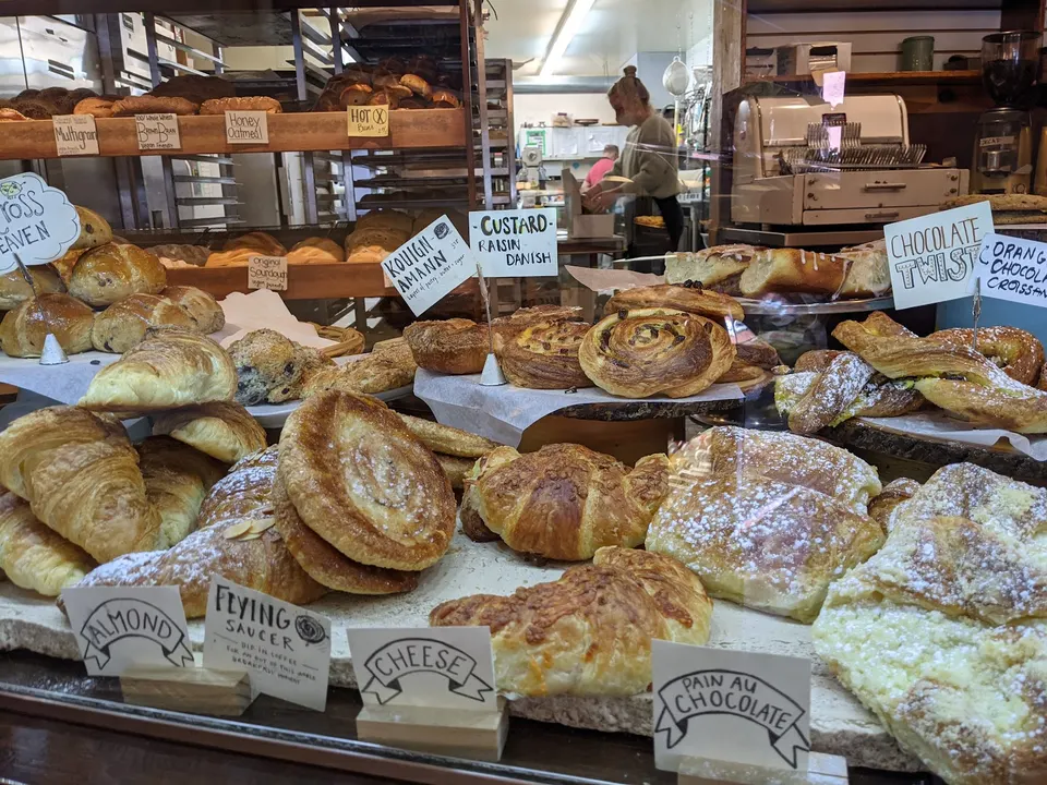 A bakery display case filled with various fresh pastries including croissants, danishes, and pain au chocolat, with hand-written labels in front.