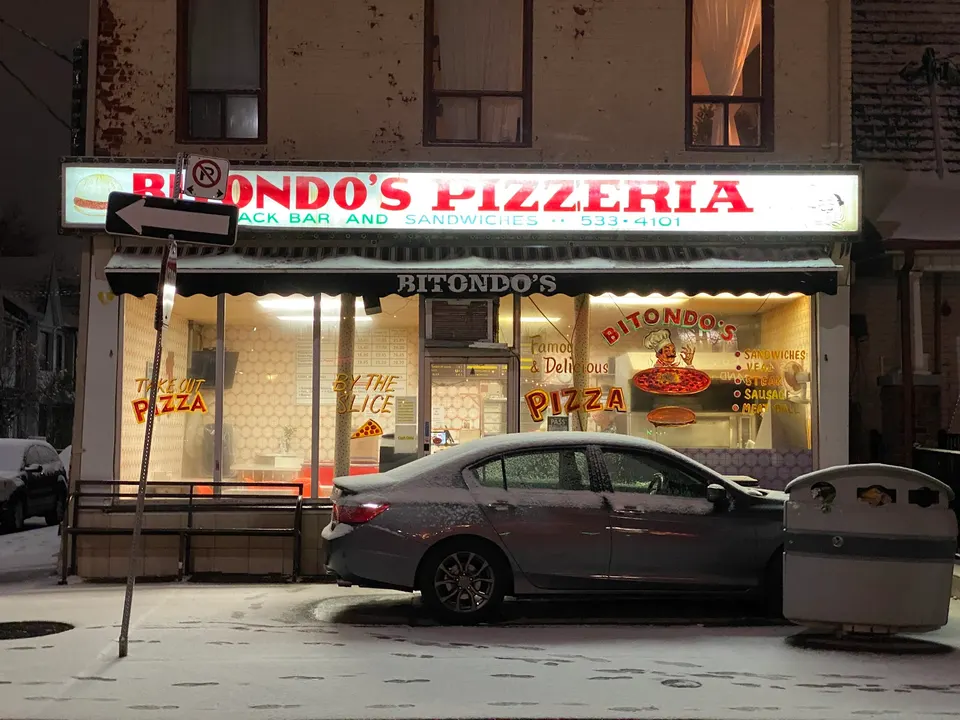 The exterior of Bitondo's Pizzeria at night, covered in snow. A gray sedan is parked in front of the brightly lit storefront window, which displays pizza signs.