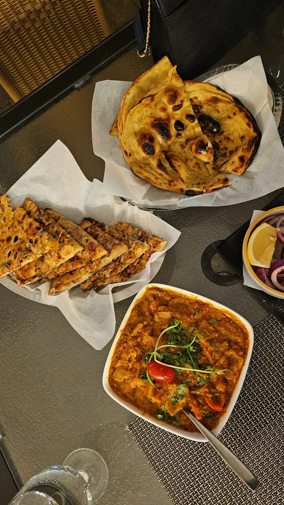 An overhead view of a table set with Indian dishes, featuring a bowl of rich orange curry garnished with herbs and a cherry tomato, a basket of sliced stuffed flatbread, and a basket of tandoori roti.