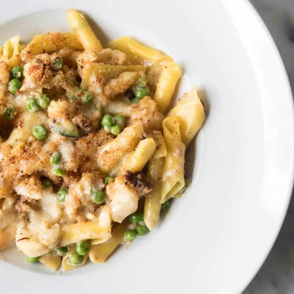 A close-up, overhead shot of a white bowl filled with penne pasta tossed in a creamy white sauce, green peas, and topped with toasted breadcrumbs.