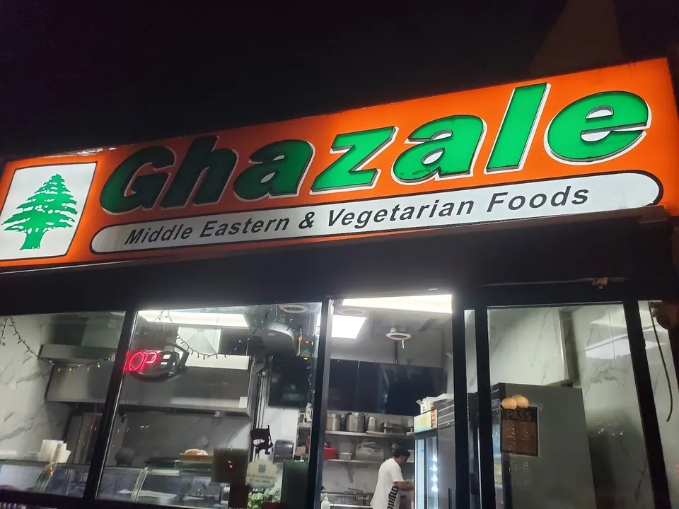Brightly lit orange and green storefront sign for Ghazale Middle Eastern & Vegetarian Foods at night, showing the interior kitchen through the window.